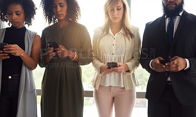 Buy stock photo Cropped shot of a group of young businesspeople using their cellphones while standing in a line in their office