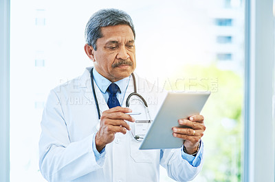 Buy stock photo Shot of an focused mature male doctor browsing on a digital tablet while standing inside of a hospital during the day