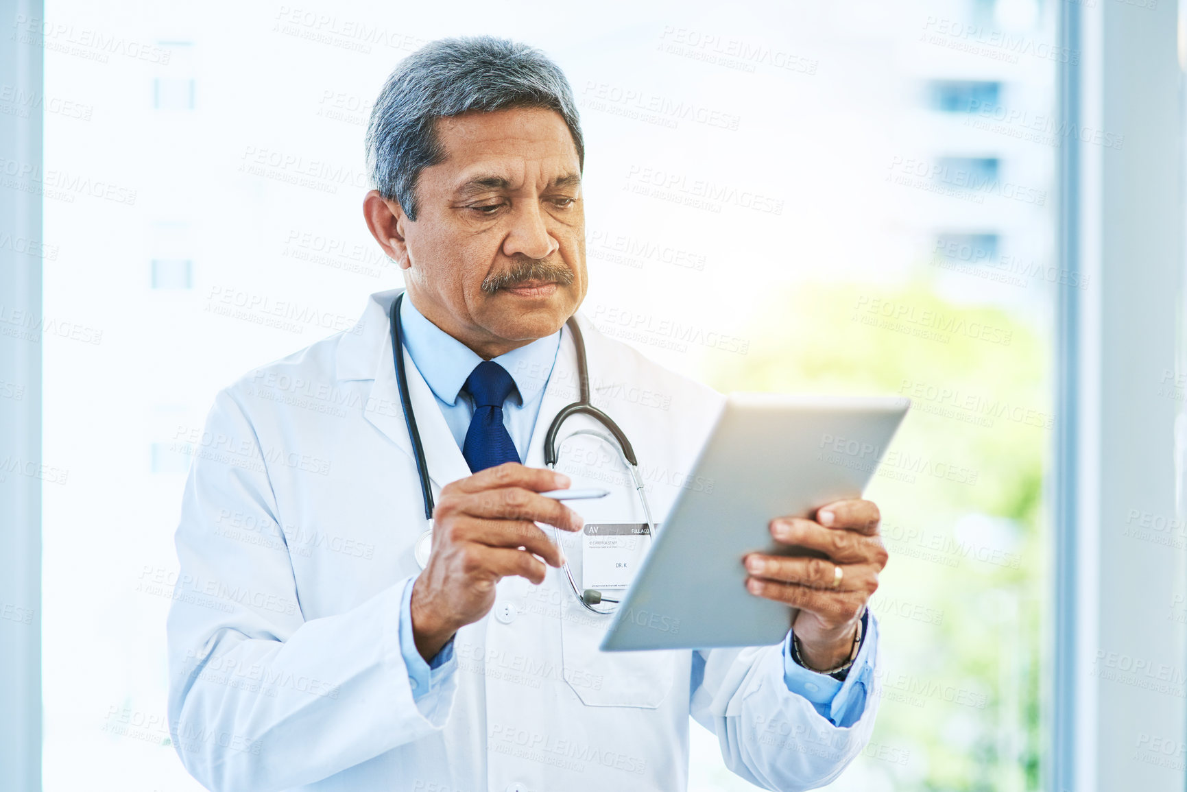 Buy stock photo Shot of an focused mature male doctor browsing on a digital tablet while standing inside of a hospital during the day