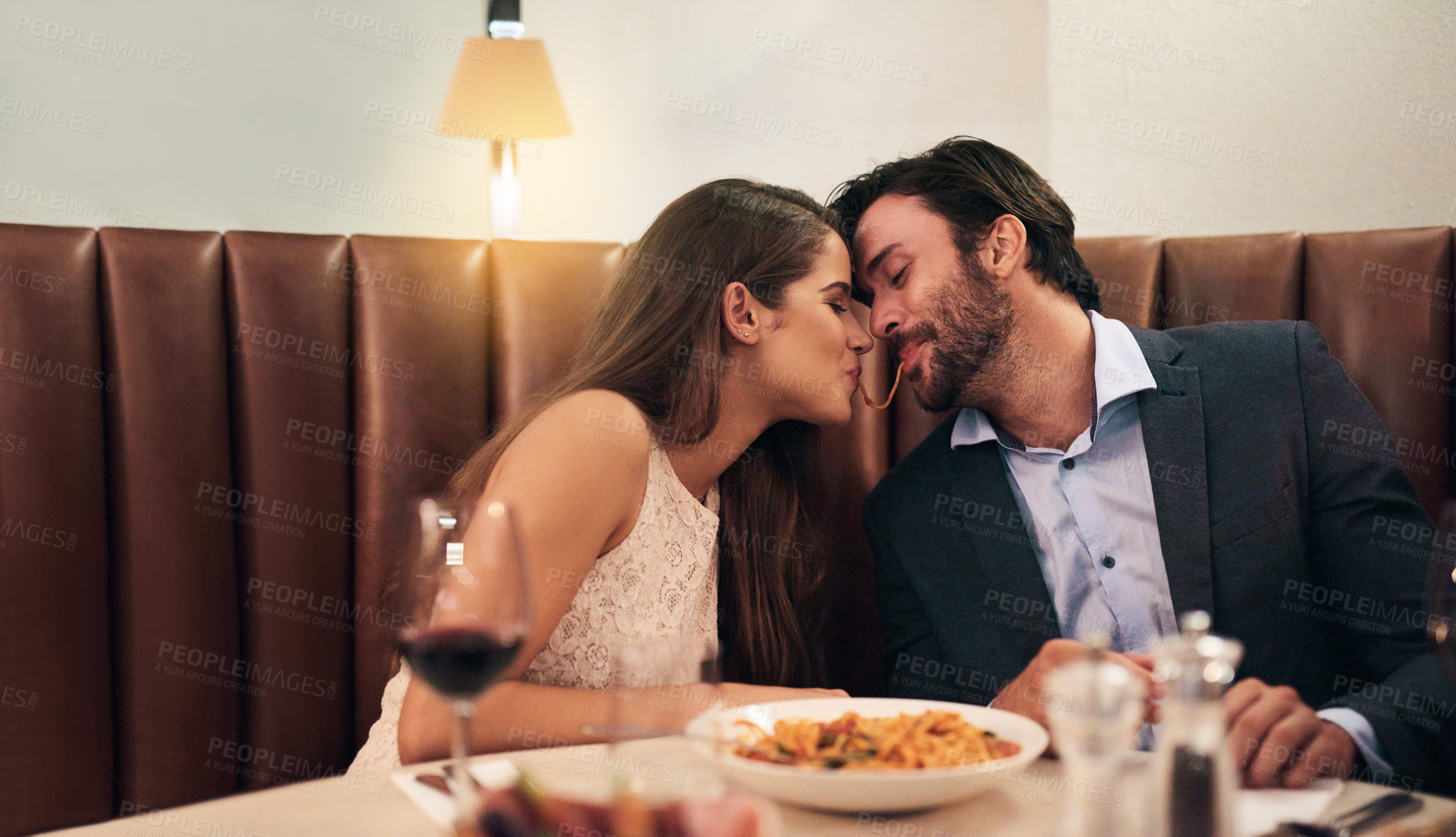 Buy stock photo Happy couple, sharing food and kiss for dinner date, embrace or relationship romance at night in restaurant. Man and woman lean with noodle mouth for intimate kissing in fine dining on valentines day