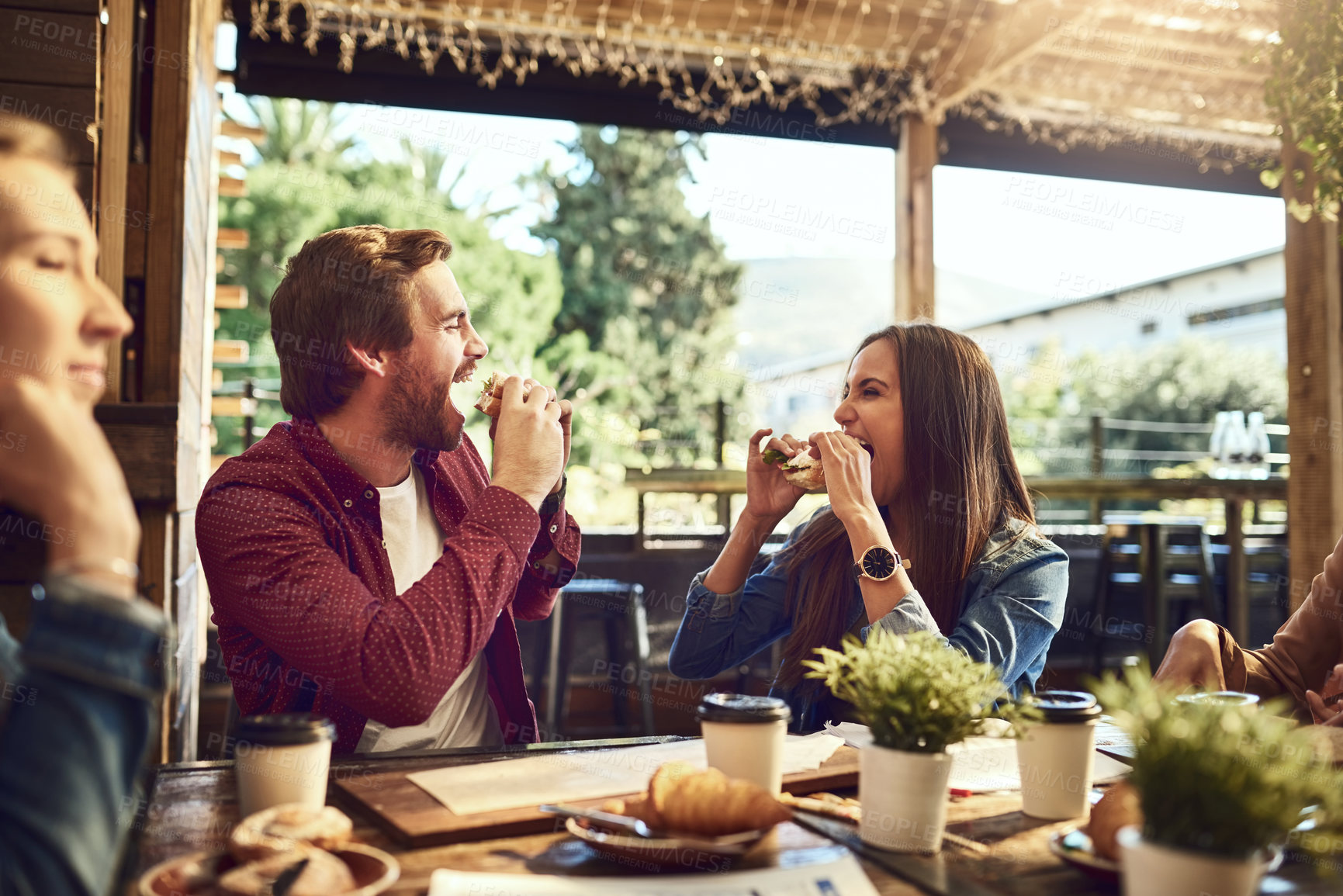 Buy stock photo Cropped shot of an affectionate young couple biting into their sandwiches while out for lunch at a cafe with friends