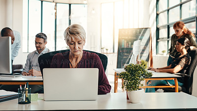 Buy stock photo Shot of a mature businesswoman using a laptop in a modern office with her colleagues in the background