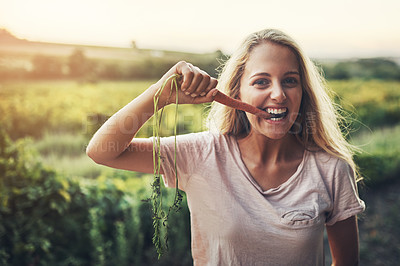 Buy stock photo Cropped portrait of a young woman holding up a carrot and taking a bite with her farmland in the background