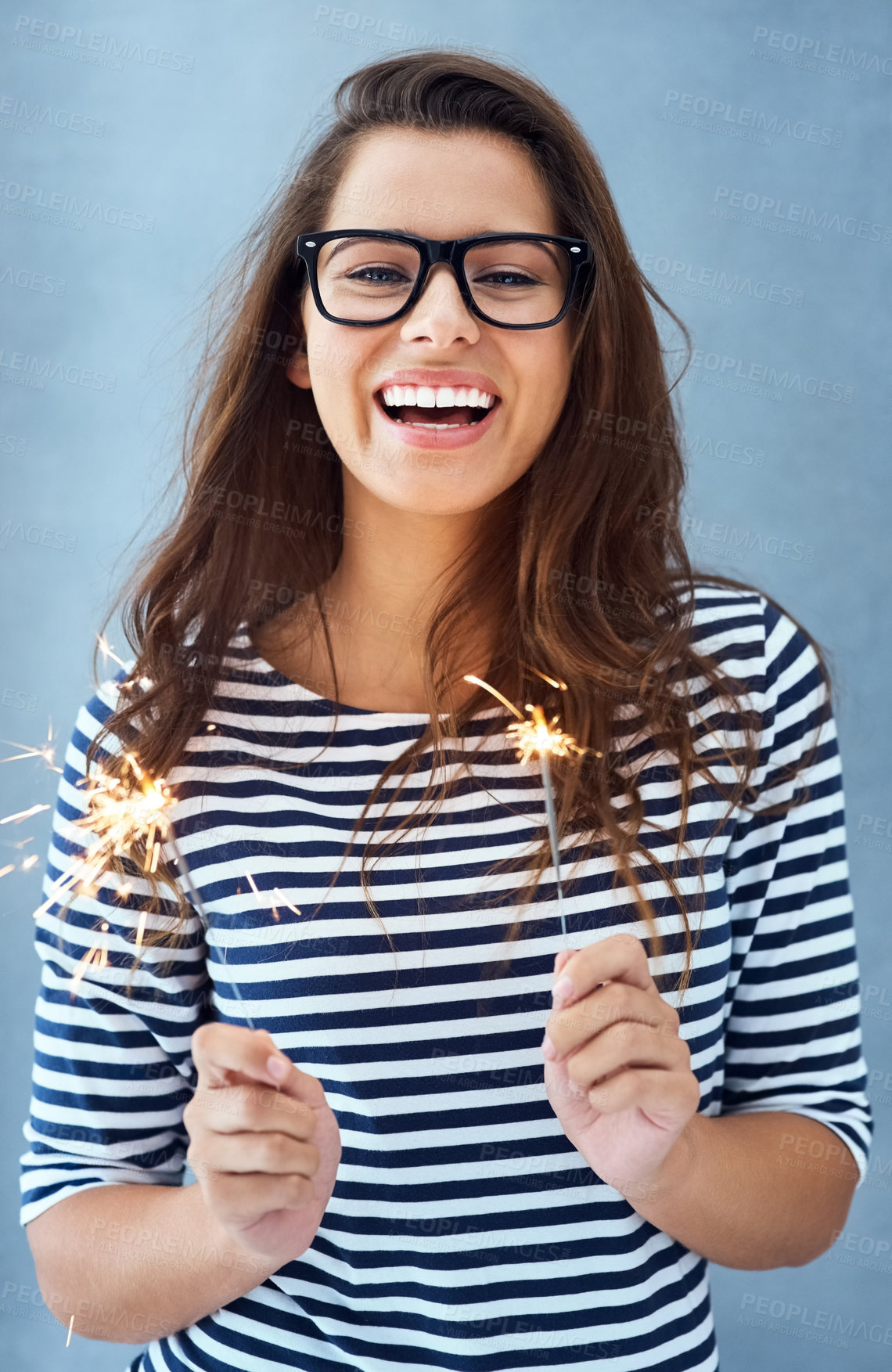 Buy stock photo Portrait, woman and happy in studio with sparklers for celebration isolated on blue background with light. Glasses, smile and fun event with birthday or new year with glow, vision and fireworks.