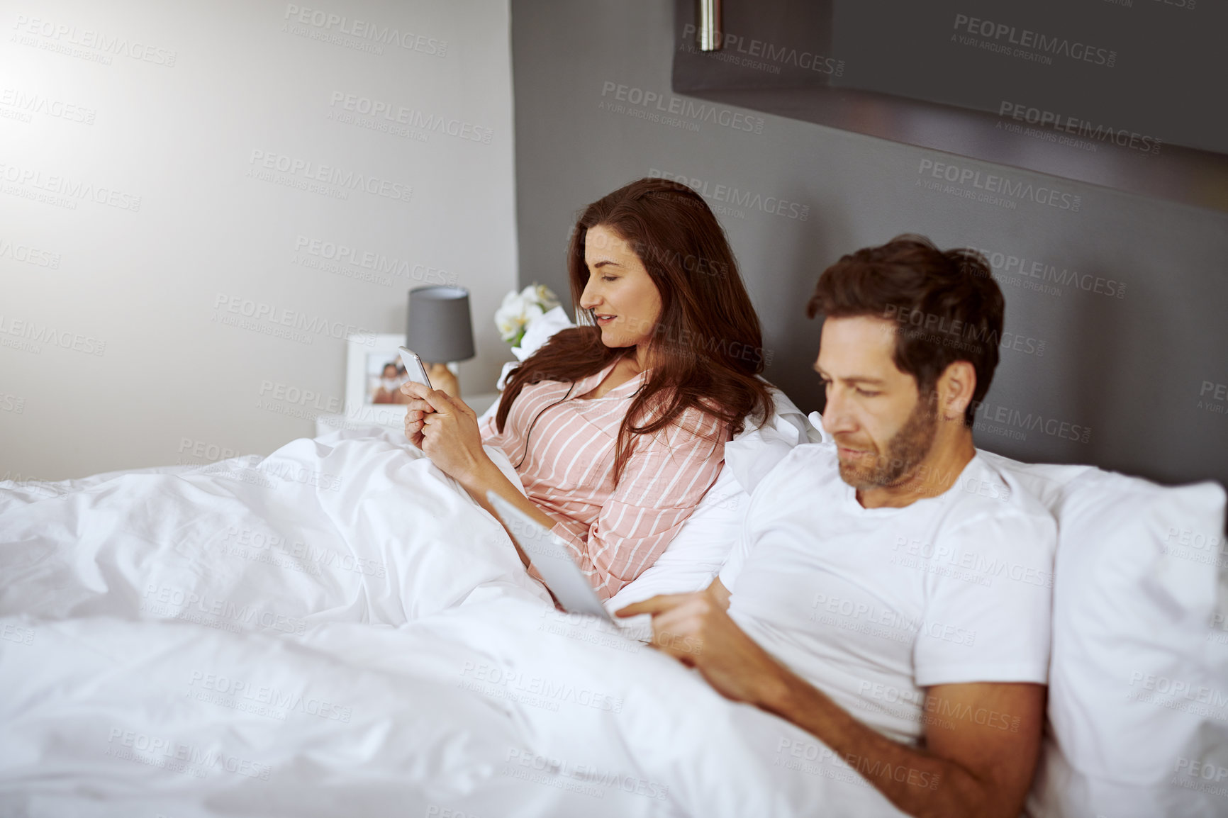 Buy stock photo Cellphone, tablet and couple in bed in home scrolling, browsing and networking on social media in morning. Rest, digital technology and phone with man and woman relaxing in bedroom at house.