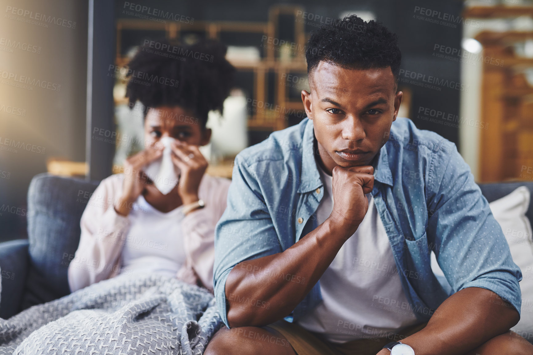 Buy stock photo Shot of a young woman blowing her nose with her husband sitting next to her on the sofa