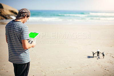 Buy stock photo Shot of a focused young man holding a remote control to fly a drone on a beach outside during the day