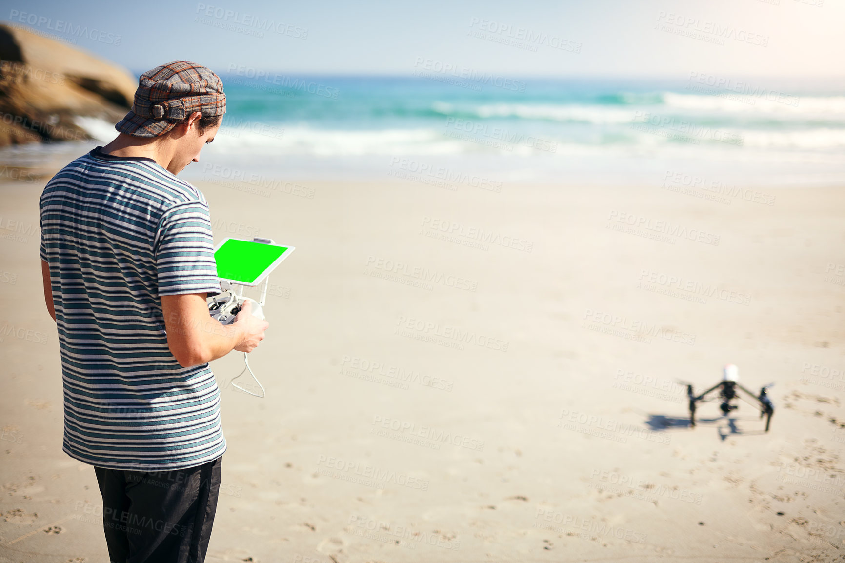 Buy stock photo Shot of a focused young man holding a remote control to fly a drone on a beach outside during the day