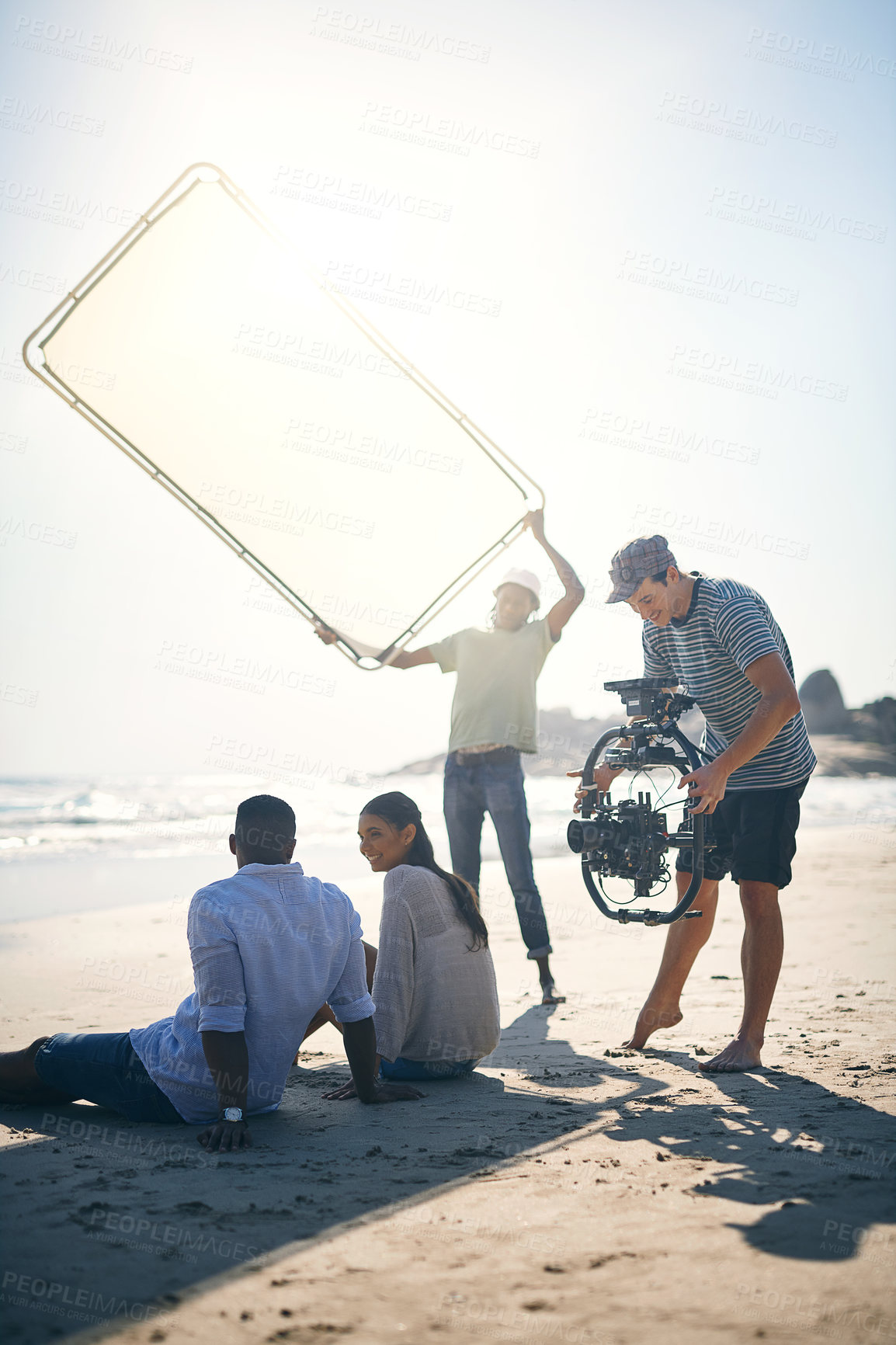 Buy stock photo Shot of a man holding up a sun screen to dim the sun rays on two models that are being filmed by a videographer