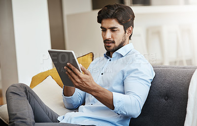 Buy stock photo Cropped shot of a handsome young man using his tablet while sitting on his sofa in the living room at home