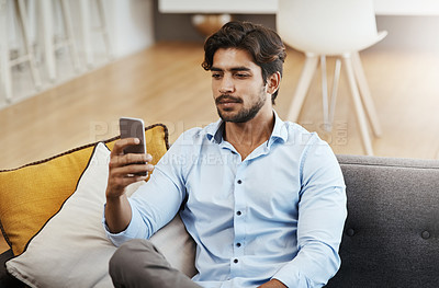 Buy stock photo Cropped shot of a handsome young man sending a text while sitting on his sofa in the living room at home