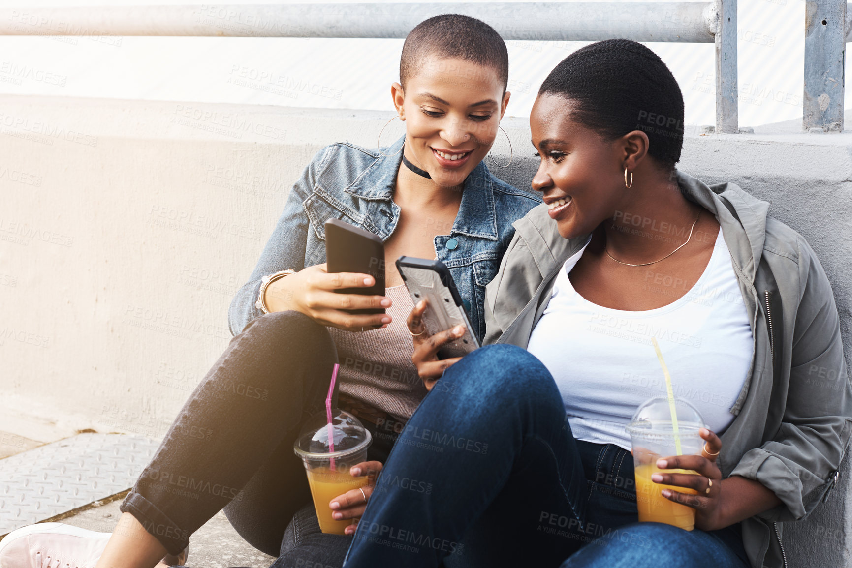 Buy stock photo Shot of two young women in the city sitting down while laughing and holding their drinks reading through text messages