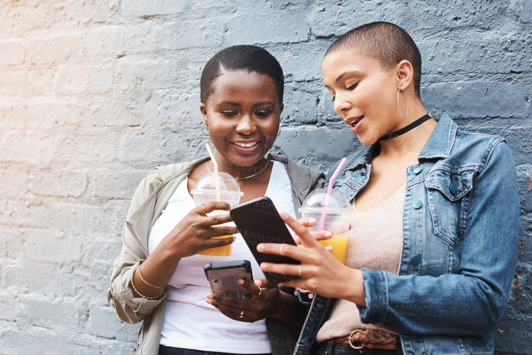 Buy stock photo Shot of two young women standing beside a building smiling and reading through text messages while holding their cool drinks