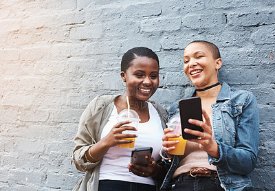 Buy stock photo Shot of two young women standing beside a building smiling and reading through text messages while holding their cool drinks