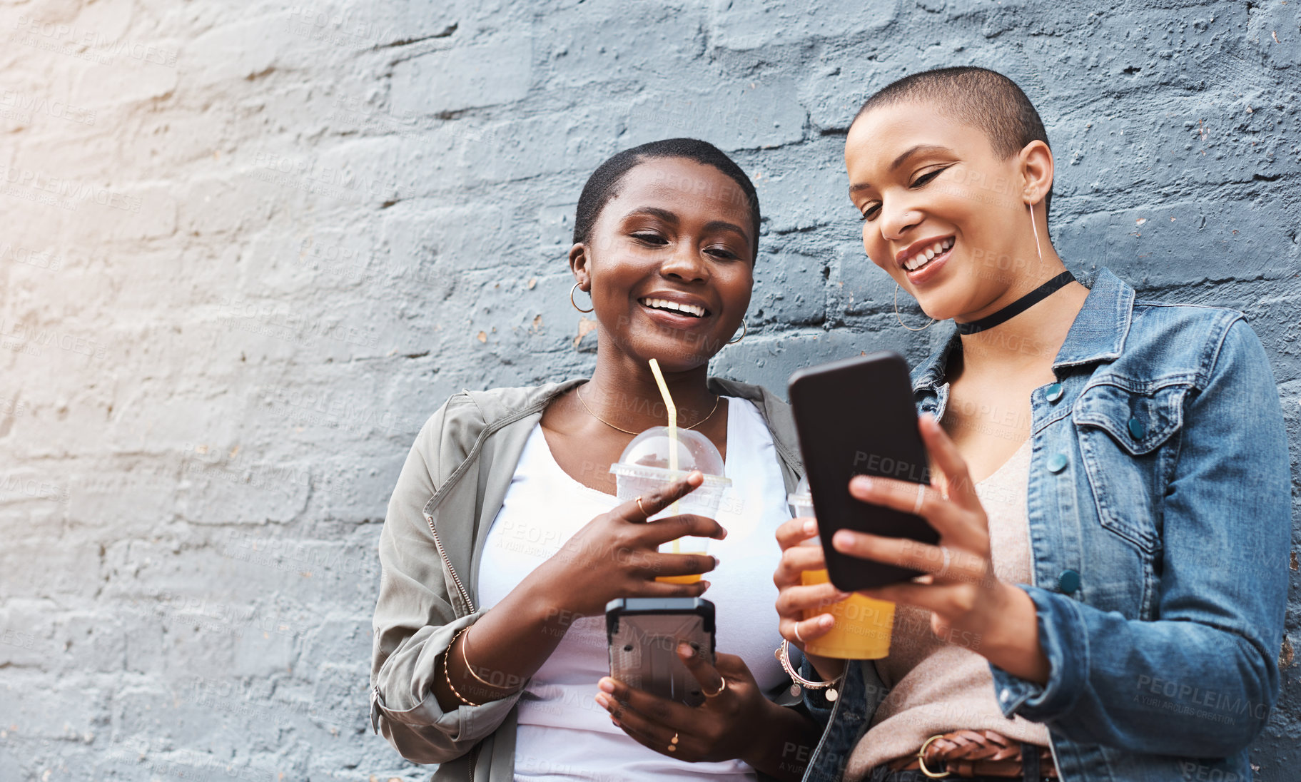 Buy stock photo Shot of two young women standing beside a building smiling and reading through text messages while holding their cool drinks