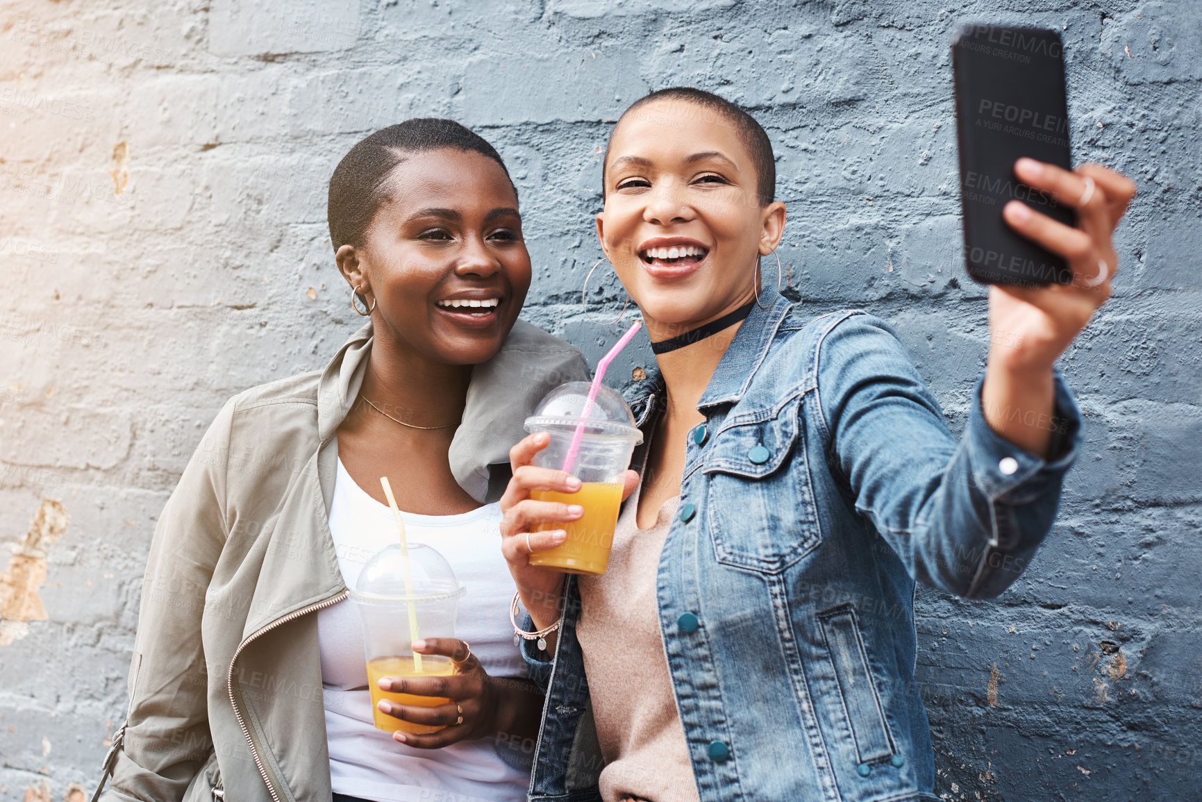 Buy stock photo Shot of two young women standing beside a building smiling and taking selfies while holding their cool drinks