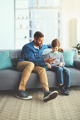 Buy stock photo Shot of a cheerful little boy and his father watching television together while being seated on the sofa at home
