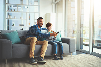 Buy stock photo Shot of a cheerful little boy and his father watching television together while being seated on the sofa at home