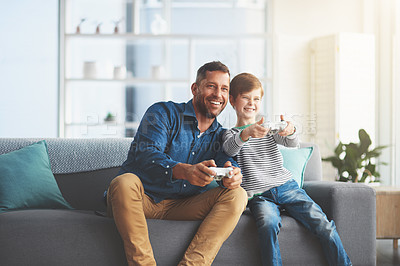 Buy stock photo Shot of a cheerful little boy and his father playing video games together on the television while being seated on the sofa at home during the day