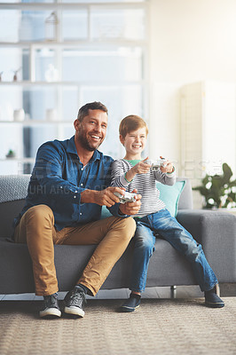 Buy stock photo Shot of a cheerful little boy and his father playing video games together on the television while being seated on the sofa at home during the day