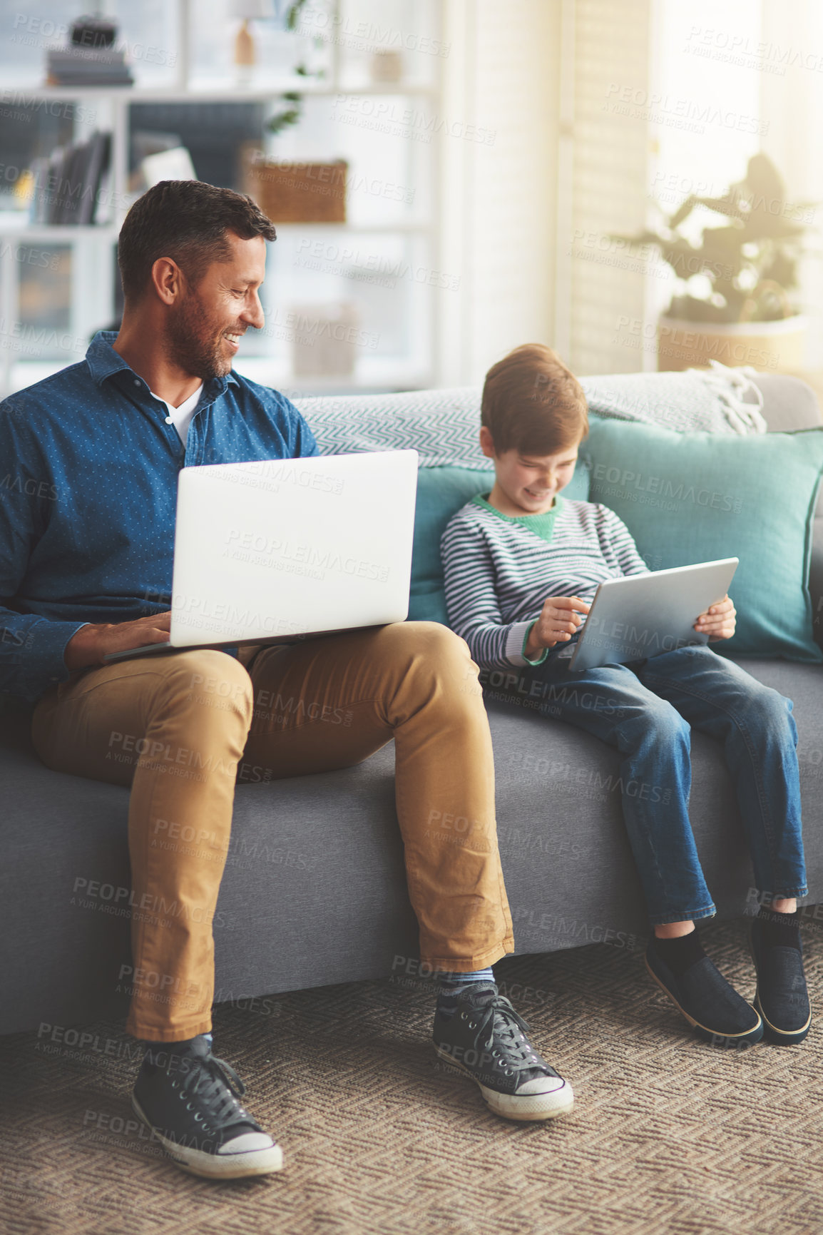 Buy stock photo Shot of a cheerful little boy and his father using a laptop and a digital tablet while being seated on a sofa at home during the day