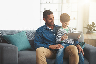 Buy stock photo Shot of a cheerful little boy browsing on a digital tablet while being seated on his father's lap at home during the day