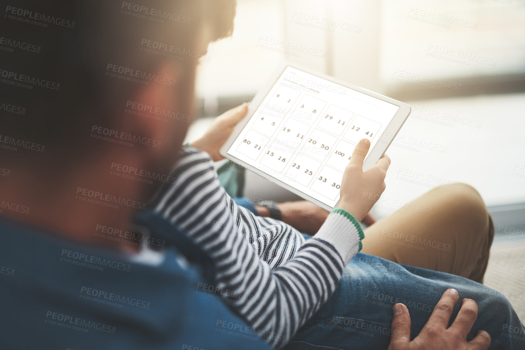 Buy stock photo Over the shoulder shot of a cheerful little boy browsing on a digital tablet while being seated on his father's lap at home during the day