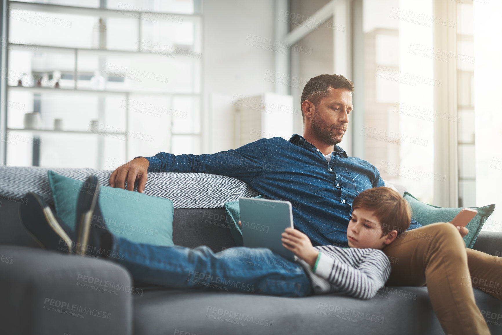 Buy stock photo Shot of a cheerful little boy browsing on a digital tablet while lying on his father's lap at home during the day