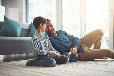 Buy stock photo Shot of a cheerful little boy and his father playing video games together on the television while being seated on the floor at home during the day