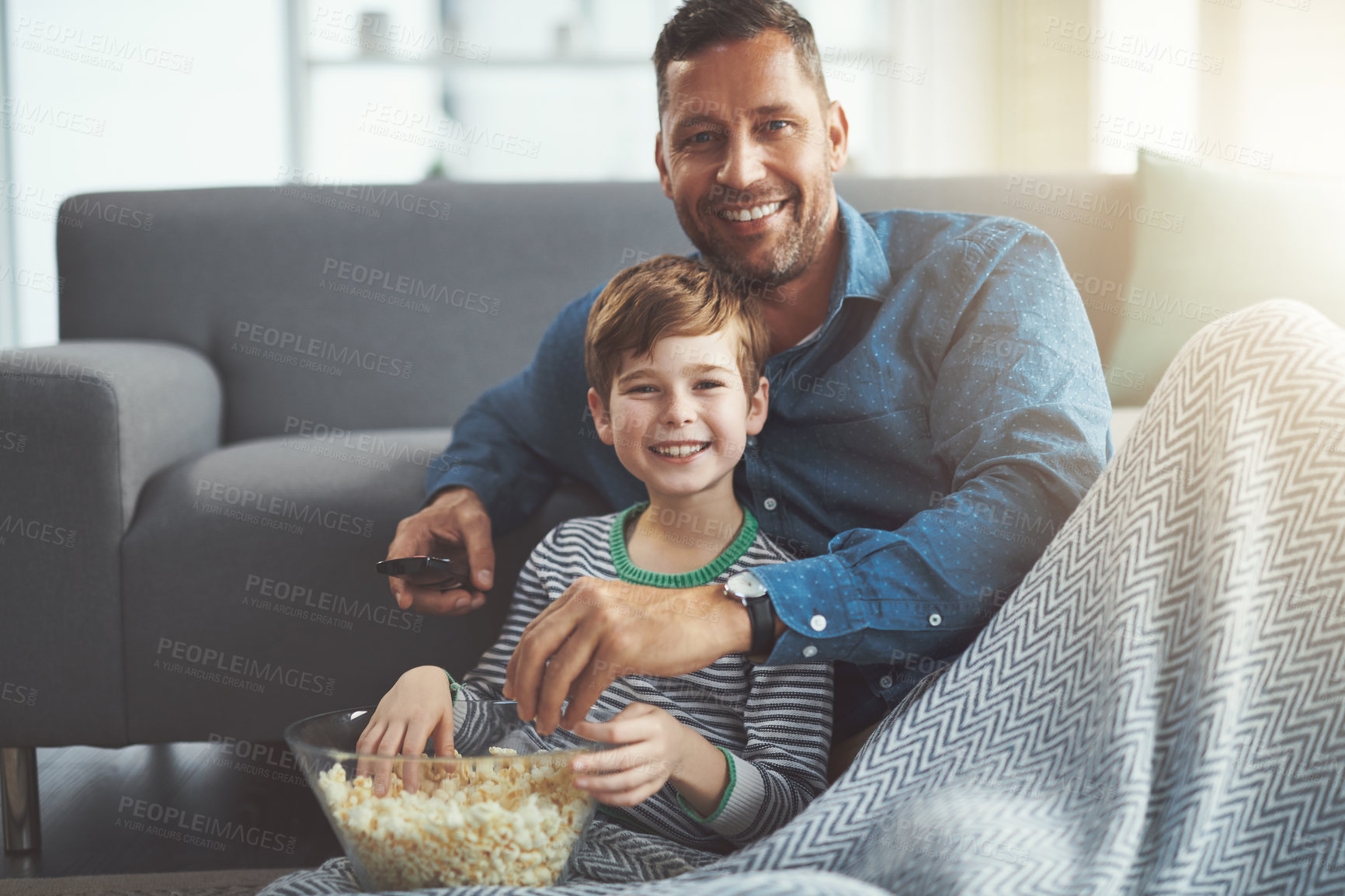 Buy stock photo Portrait of a carefree young boy and his father watching a movie together while being seated on the floor and eating popcorn at home during the day
