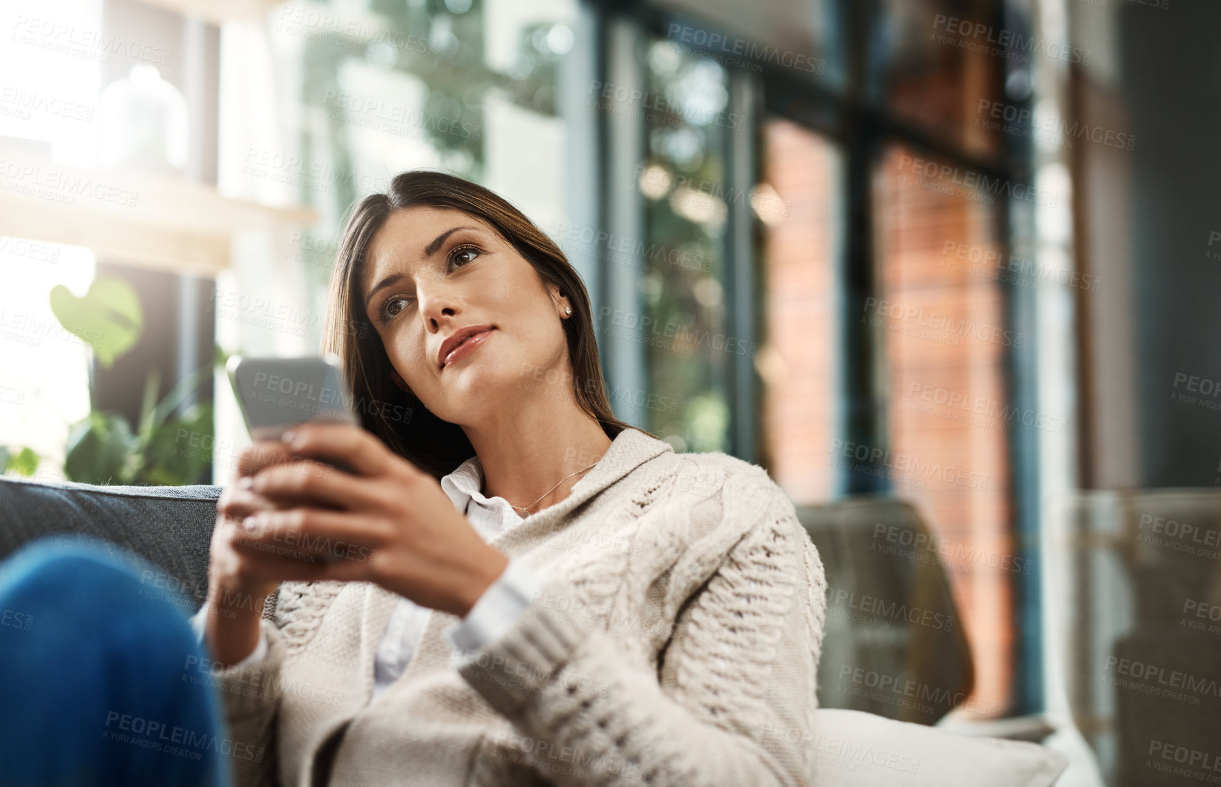 Buy stock photo Shot of an attractive young woman looking thoughtful and sending text messages while relaxing on her sofa at home