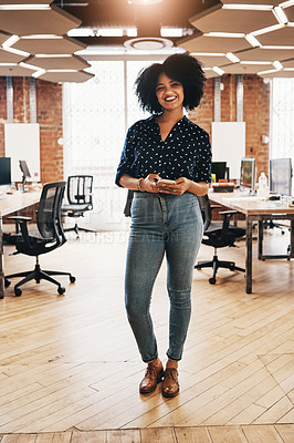 Buy stock photo Full length portrait of a cheerful young female designer holding a cellphone while standing inside of her office