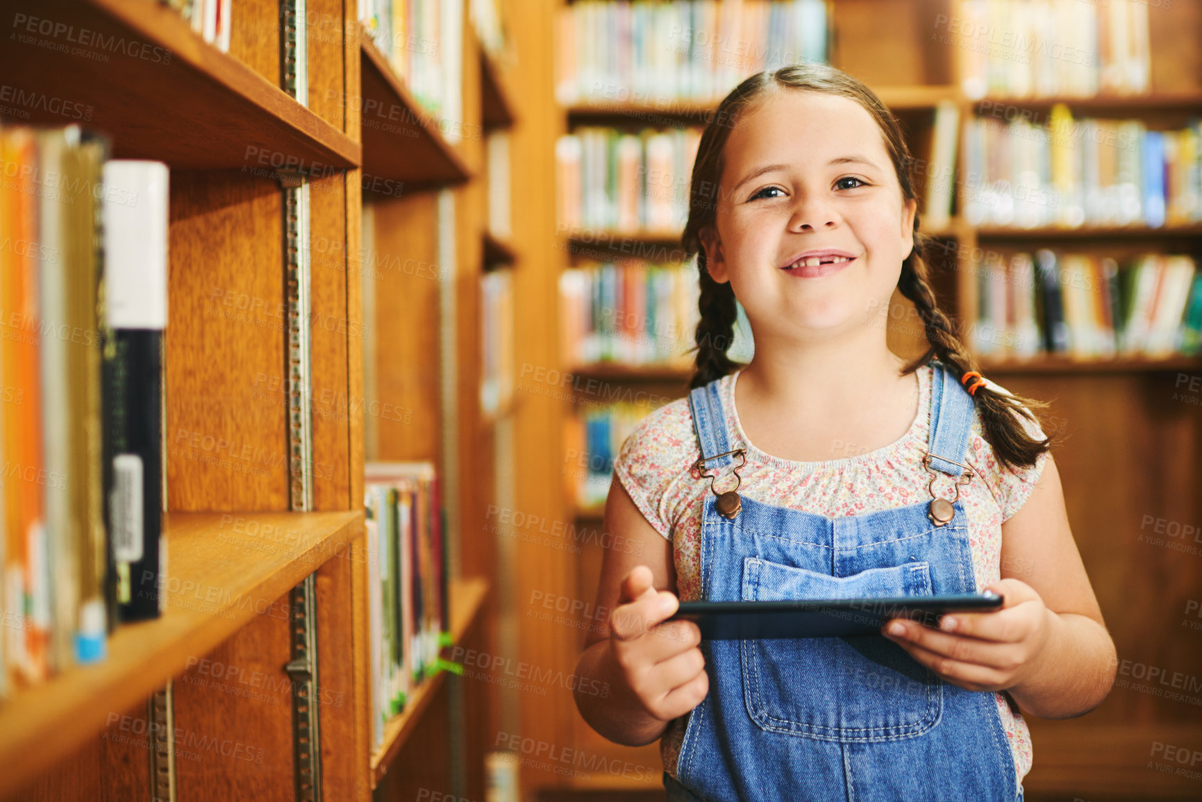 Buy stock photo Portrait of a cheerful young girl browsing on a digital tablet while standing inside of a library during the day