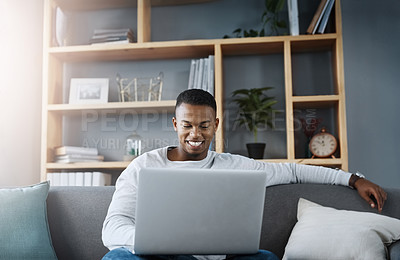 Buy stock photo Shot of a handsome young man  using his laptop while sitting on a sofa at home