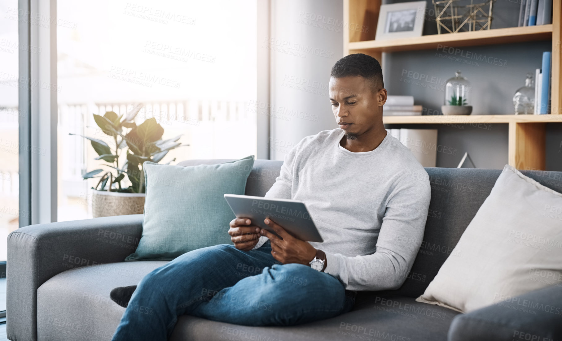 Buy stock photo Shot of a handsome young man using his digital tablet while sitting on a sofa at home