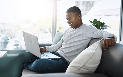 Buy stock photo Shot of a handsome young man  using his laptop while sitting on a sofa at home