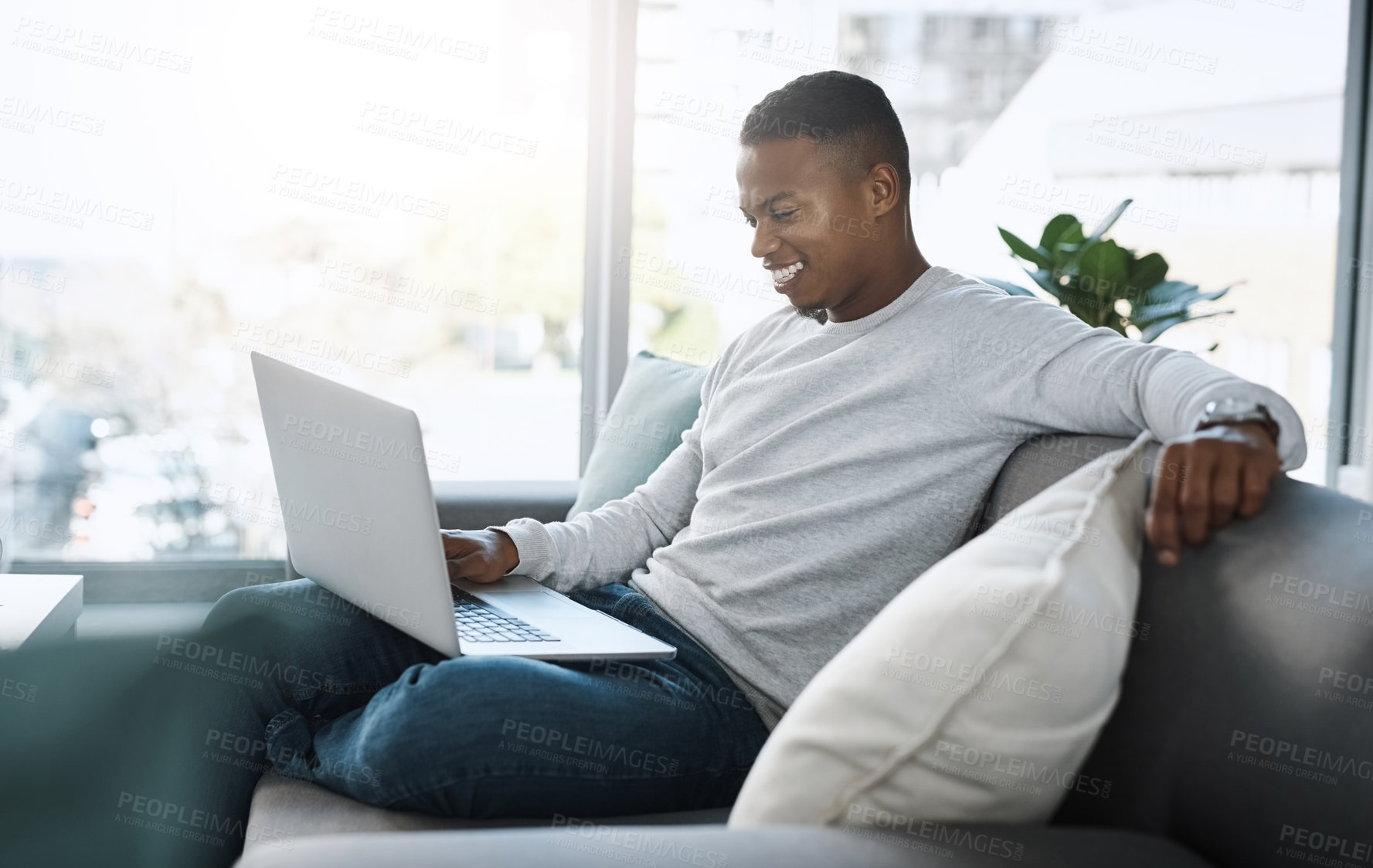 Buy stock photo Shot of a handsome young man  using his laptop while sitting on a sofa at home