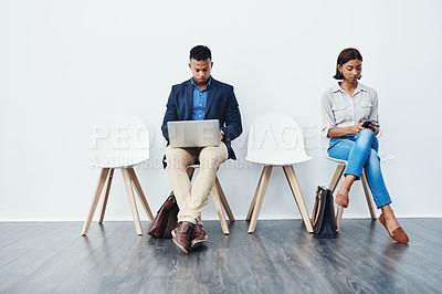 Buy stock photo Full length shot of two young businesspeople sitting down on chairs and using their tech devices against a grey background