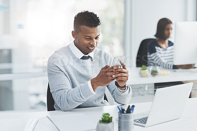 Buy stock photo Shot of a young businessman using his cellphone at his office desk with a colleague working in the background