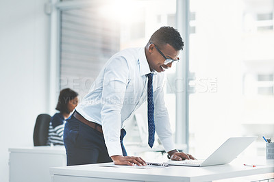 Buy stock photo Shot of a young businessman looking at figures on his laptop with a colleague of his working in the background