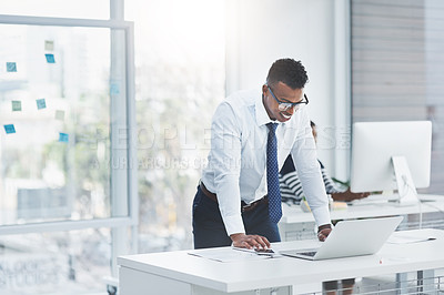 Buy stock photo Shot of a young businessman standing over his office desk and looking at figures on a laptop