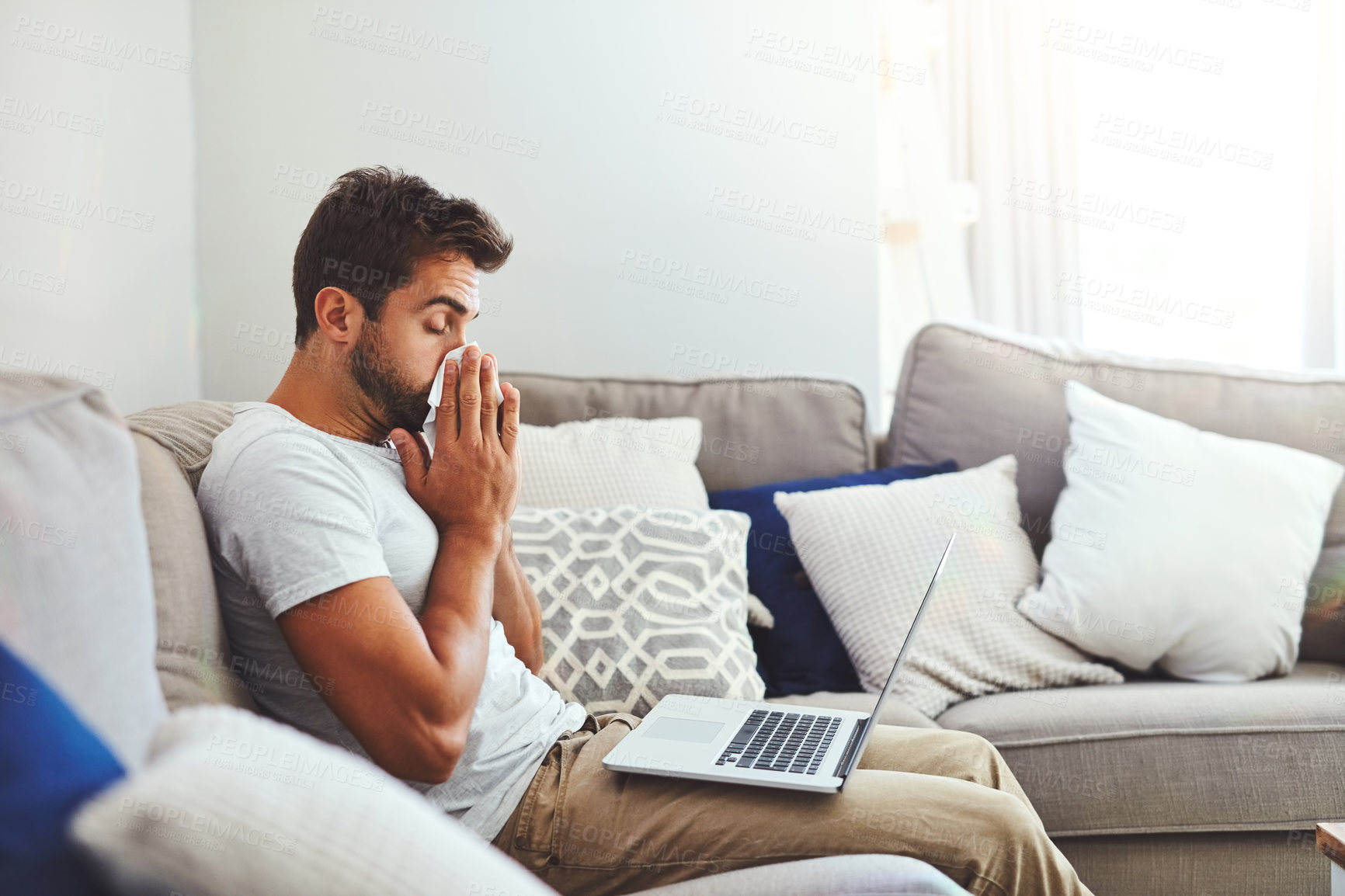 Buy stock photo Cropped shot of a handsome young man sneezing into a tissue while trying to work on his laptop at home