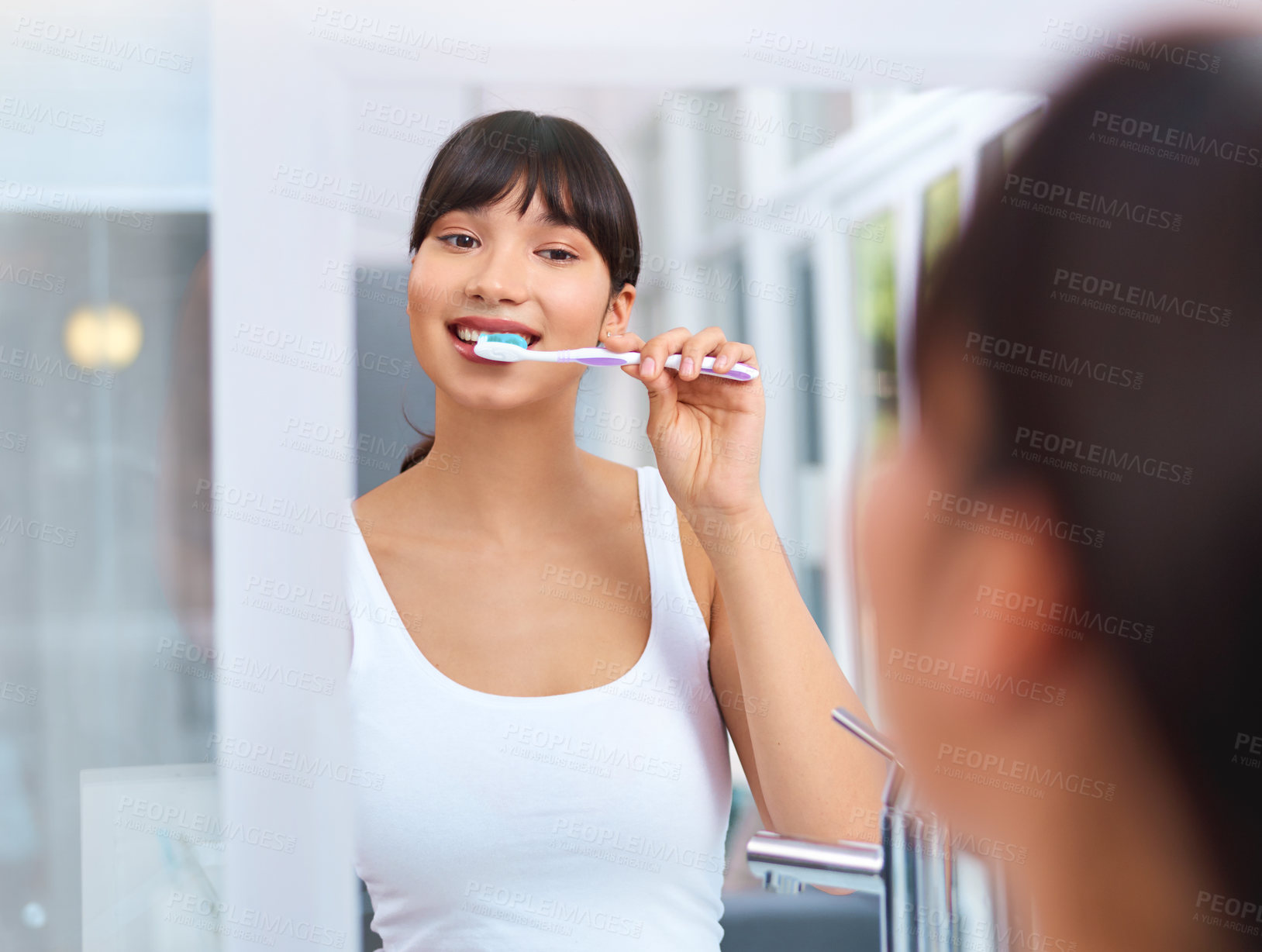 Buy stock photo Shot of a cheerful attractive young woman brushing her teeth while looking at her reflection in a mirror at home during the day
