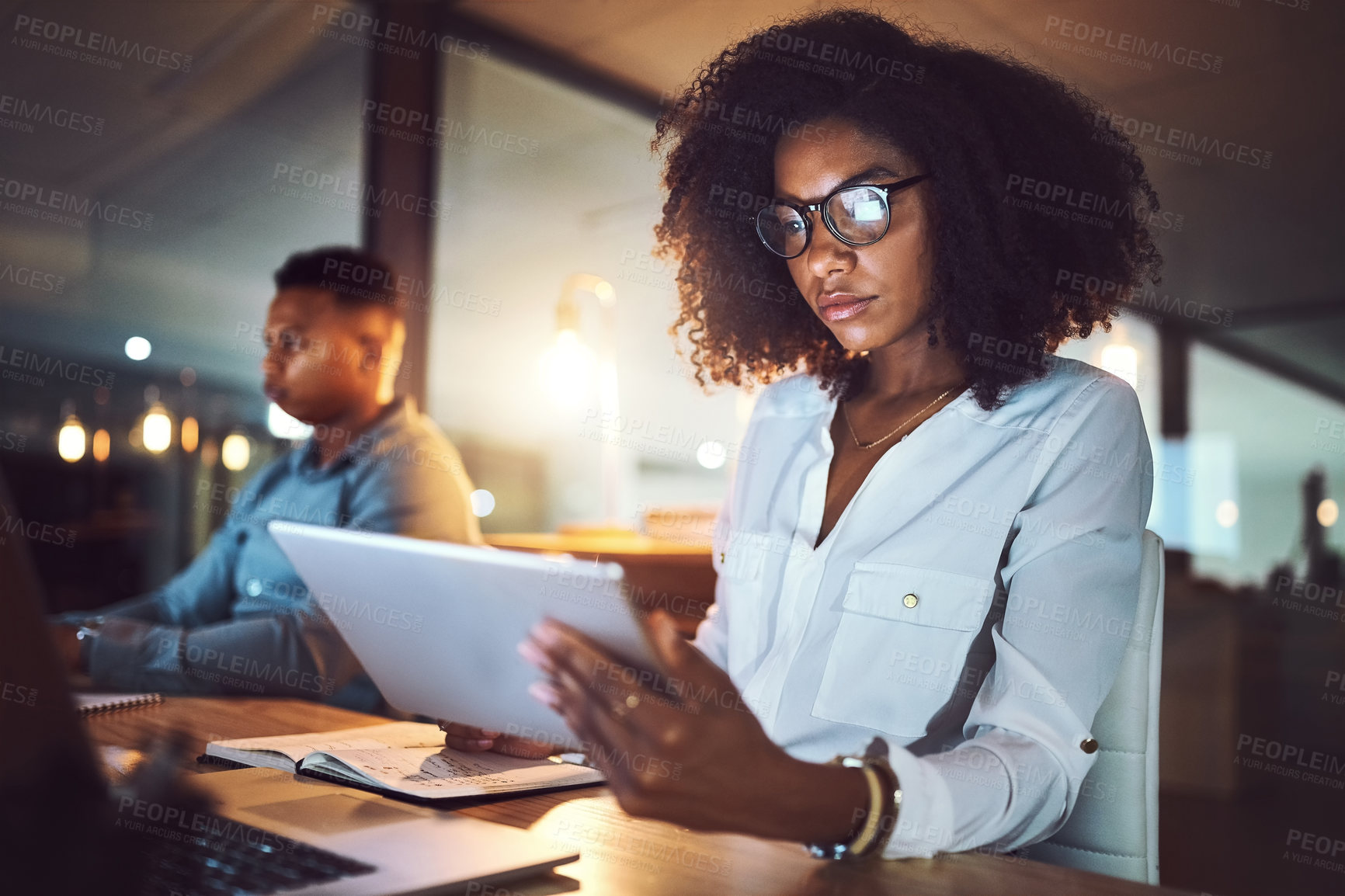 Buy stock photo Shot of a young businesswoman using a digital tablet while working alongside her colleague in an office at night