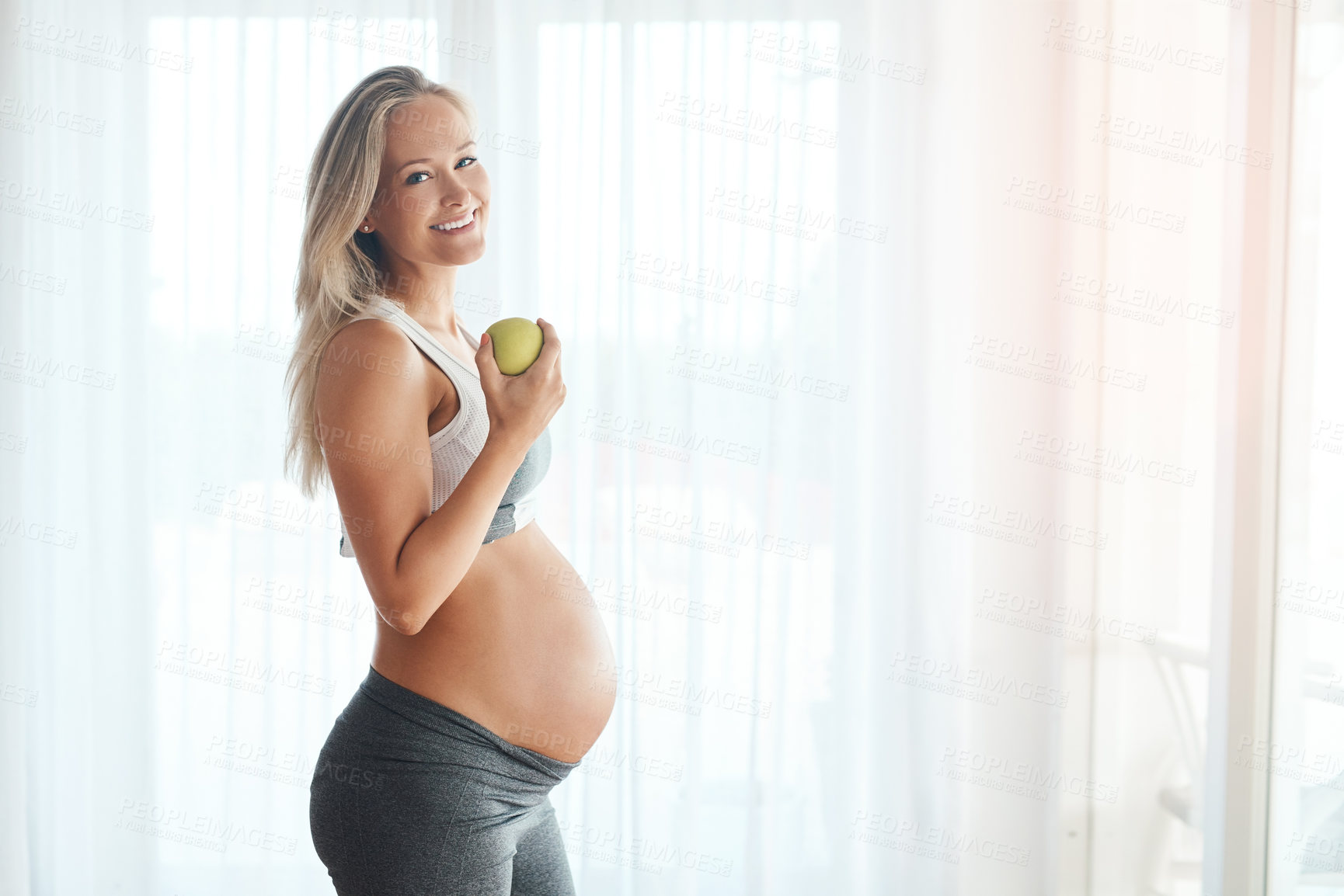 Buy stock photo Portrait of a happy pregnant woman dressed in sportswear and eating an apple at home