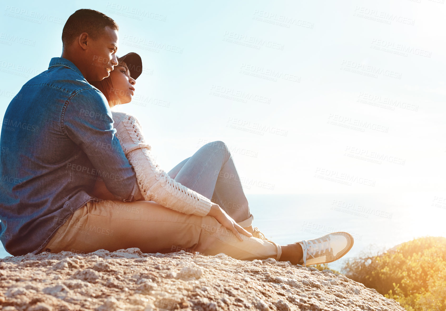 Buy stock photo Shot of a happy young couple sitting on a rock and enjoying the ocean view