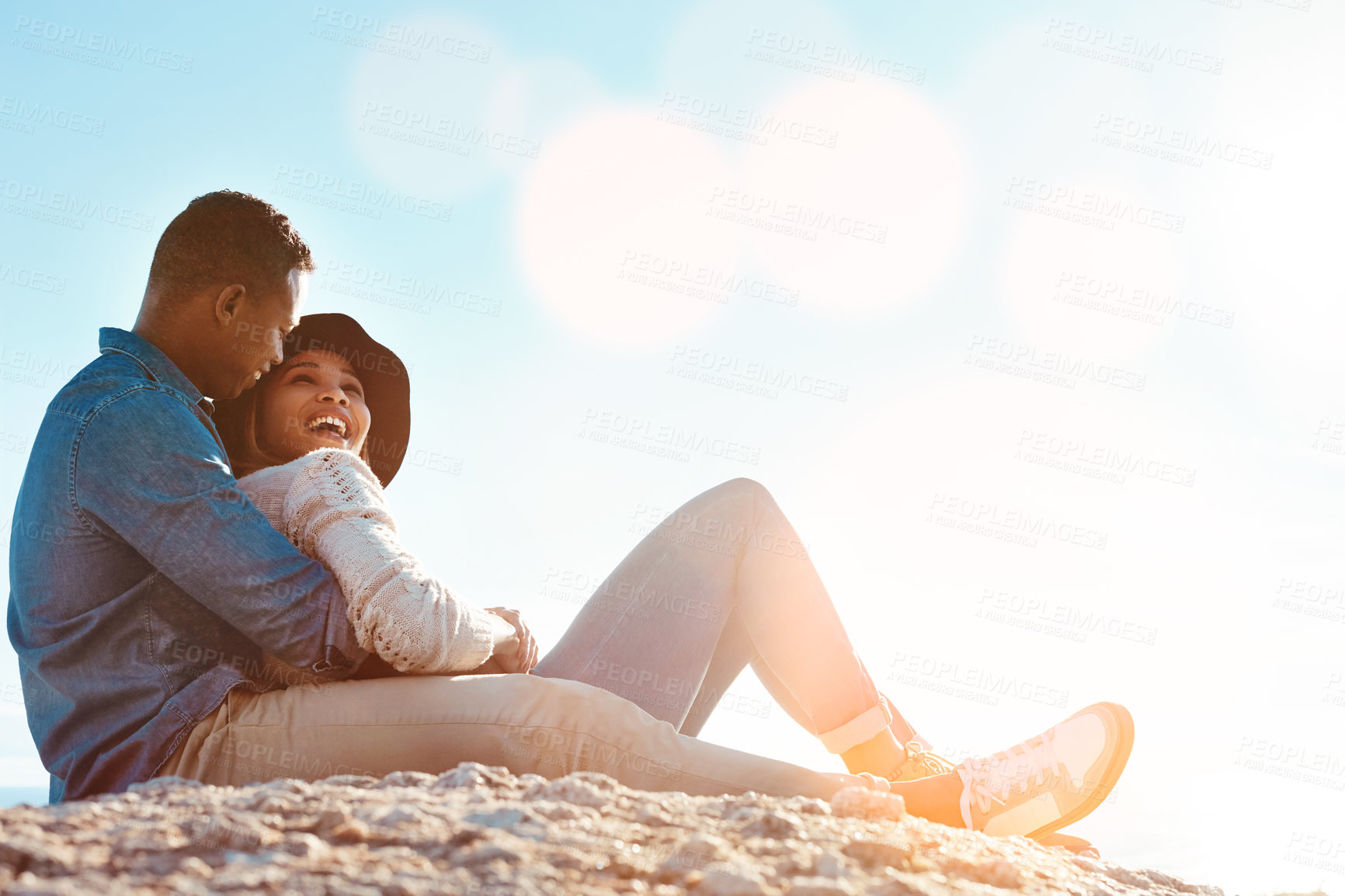 Buy stock photo Shot of a happy young couple sitting on a rock and enjoying the ocean view