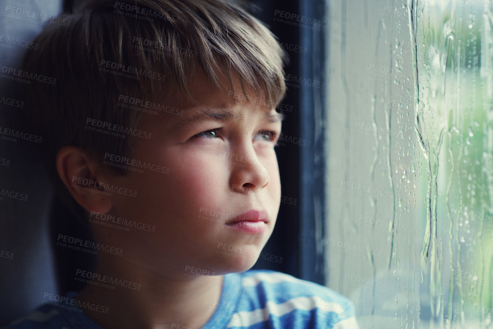 Buy stock photo Shot of a sad young boy watching the rain through a window at home
