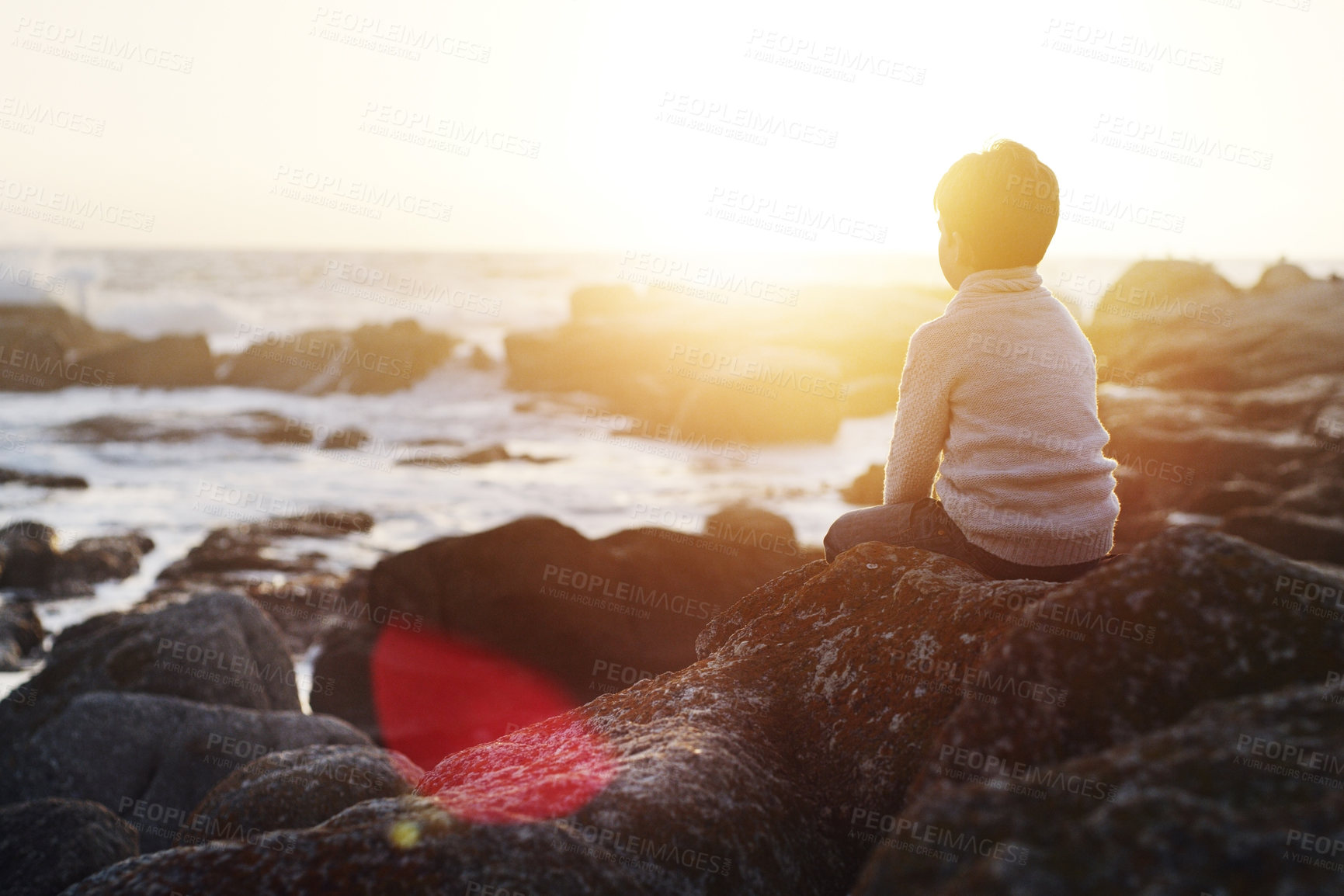 Buy stock photo Rearview shot of an adorable little boy sitting on the rocks at beach