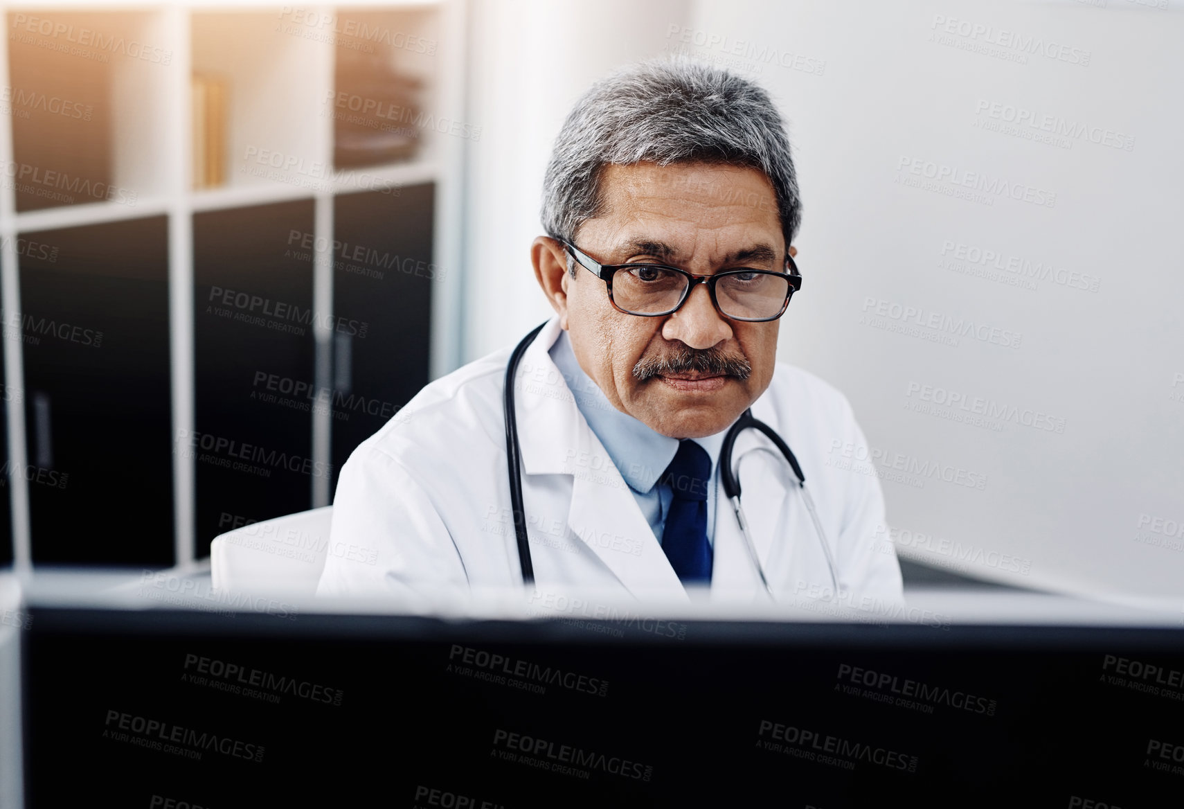 Buy stock photo Cropped shot of a focused mature male doctor working on a computer while being seated in his office during the day